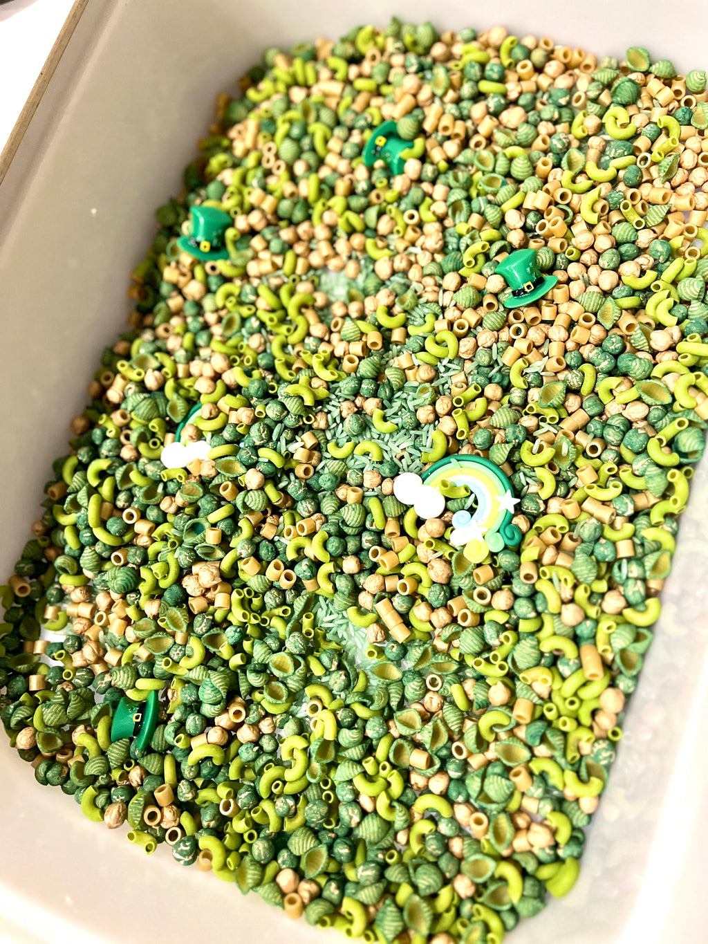 Container filled with green and beige pasta shapes, small beads, and a rainbow and green hat charms scattered inside