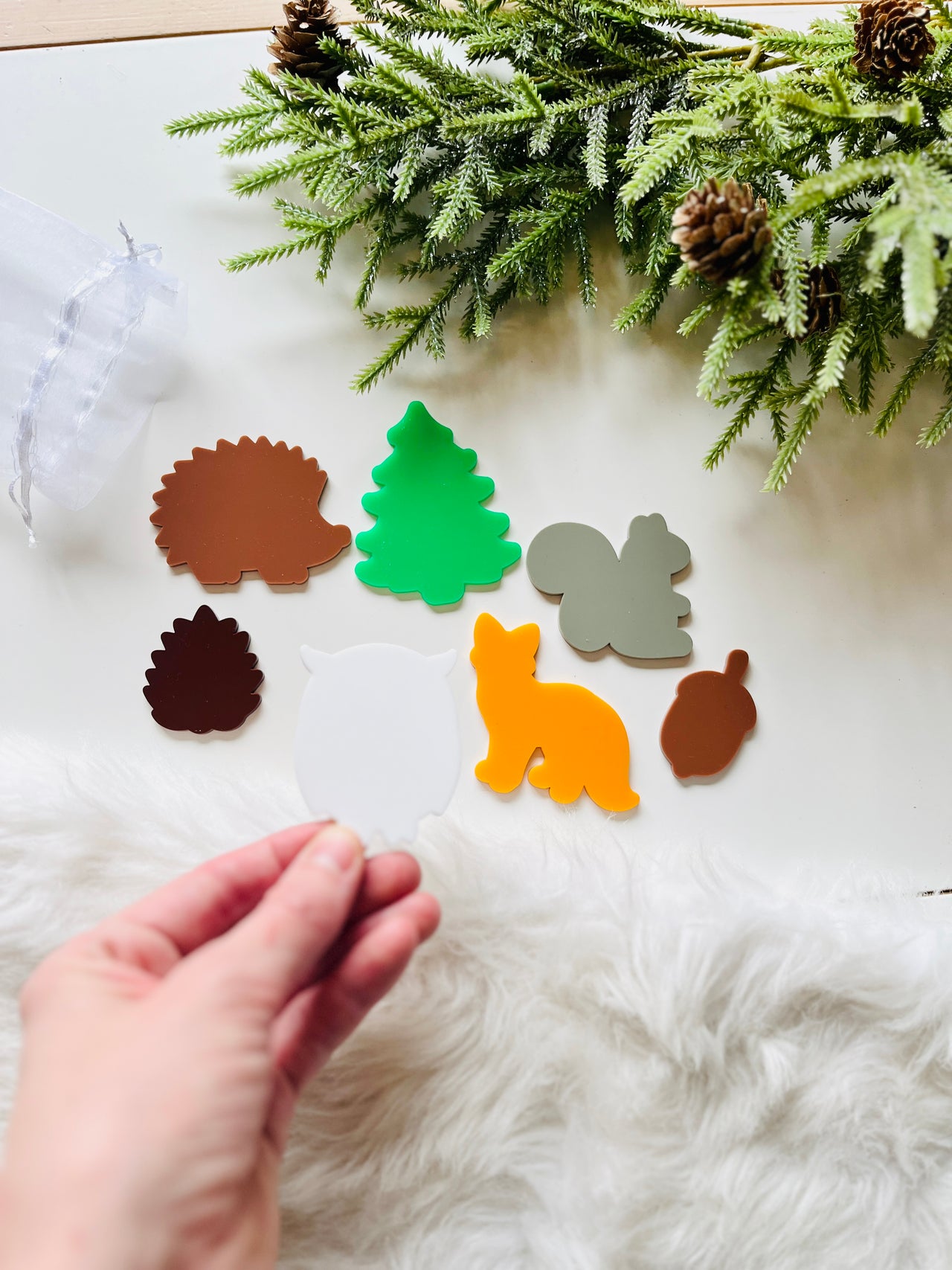 Hand holding white owl-shaped acrylic cutout with flat forest animal and tree shapes on white fur and greenery backdrop