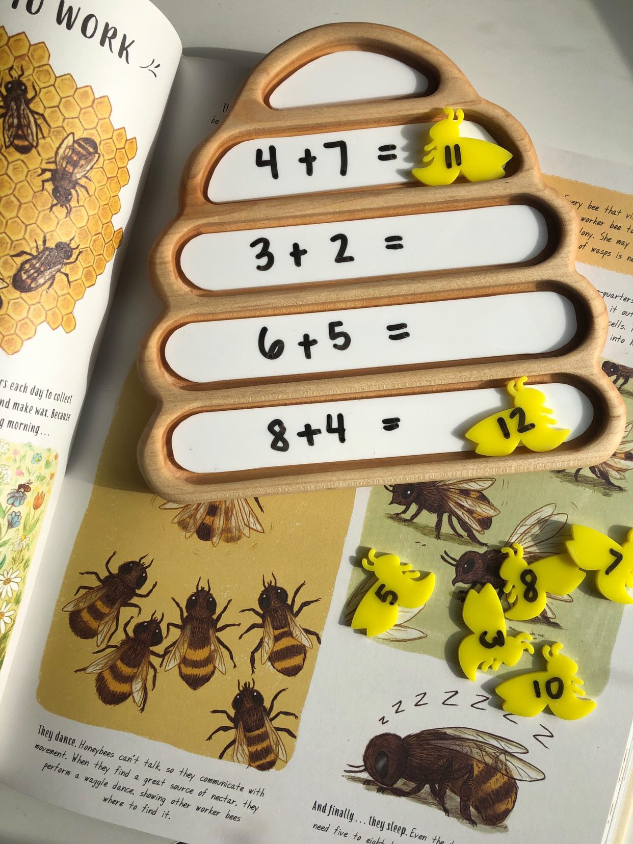 Wooden beehive-shaped board with math addition problems and yellow bee-shaped number tokens on illustrated bee book pages
