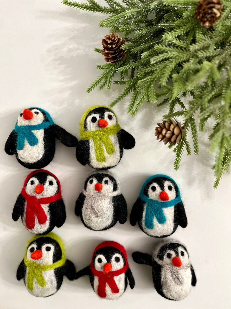 Seven handmade felt penguins with colorful scarves arranged near pine branches and cones on white background