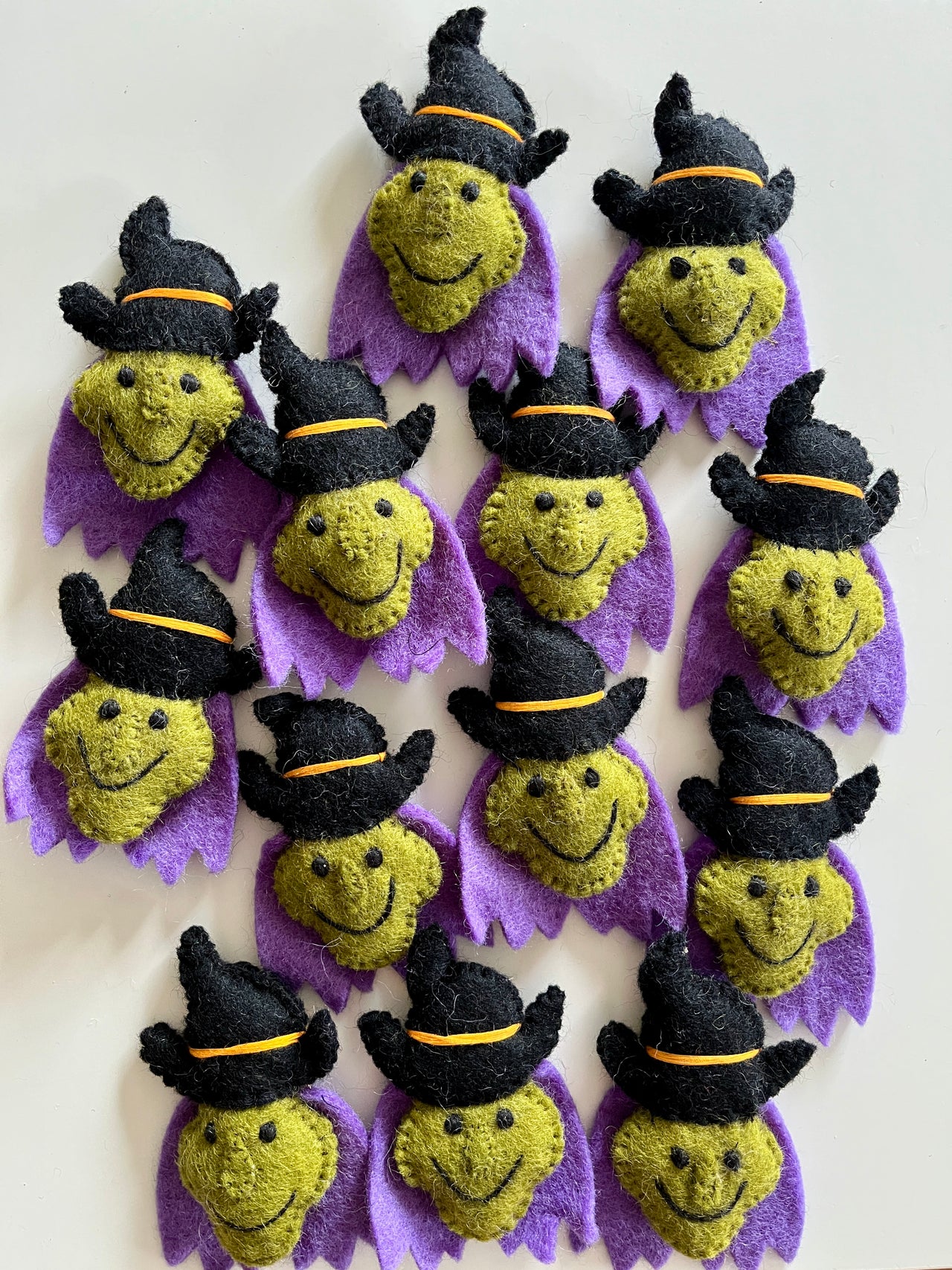 Multiple small handmade felt witch faces with green skin, black hats, and purple collars arranged on a light surface.