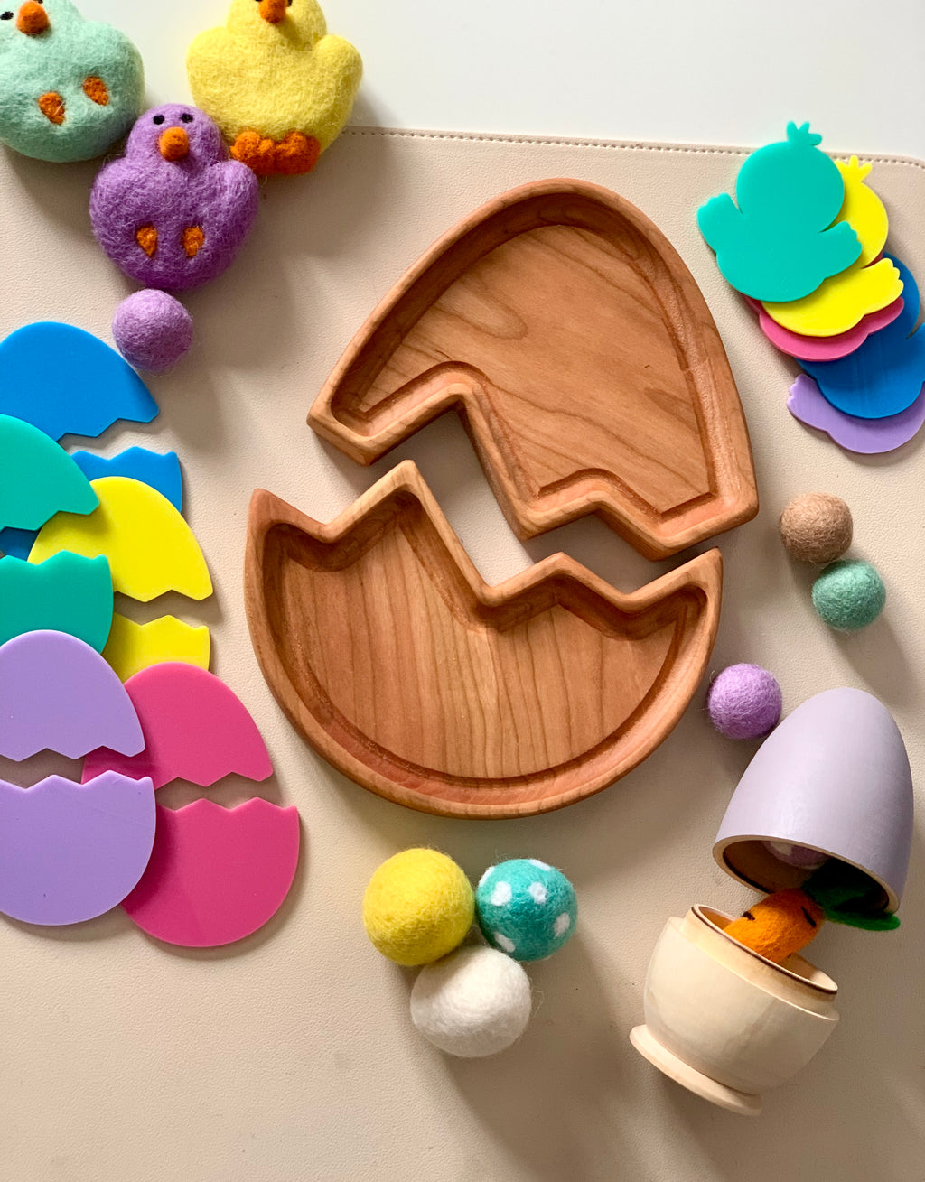 Wooden broken egg-shaped tray surrounded by colorful plastic egg pieces, felt balls, and soft chick toys on a beige surface