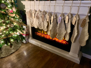 Eight cream cable knit Christmas stockings hanging on white mantle above glowing electric fireplace near decorated tree