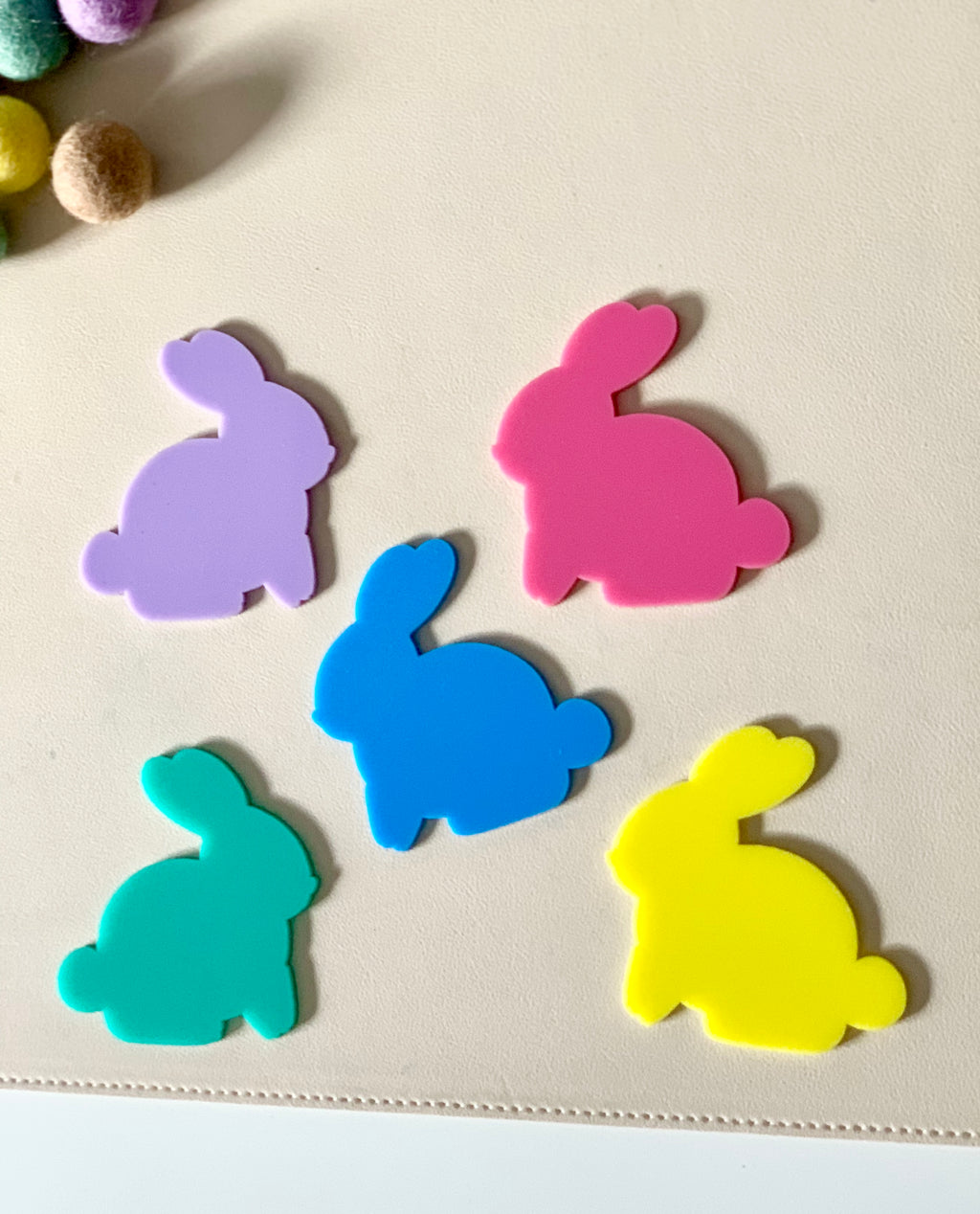 Five brightly colored bunny-shaped cutouts in purple, pink, blue, green, and yellow on a light surface near felt balls
