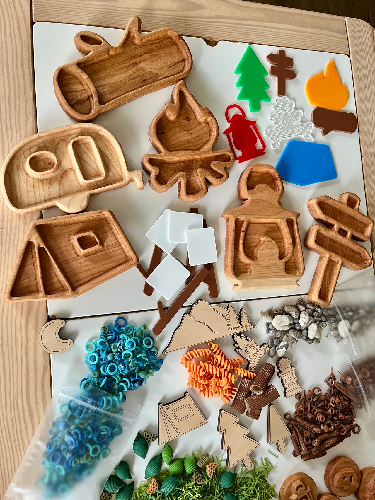 Wooden camping-themed trays including camper, campfire, lantern, tent, directional signs, and colorful nature-inspired play pieces on table