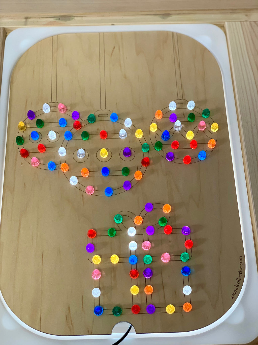 Wooden Flisat insert with engraved Christmas ball and gift shapes outlined by colorful light pegs on wooden table