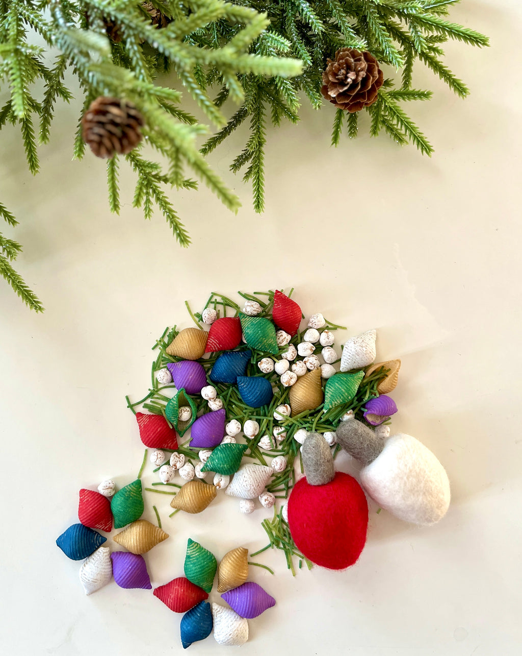 Assorted colorful fabric bulbs and white berries arranged on a white surface with pine branches and cones above
