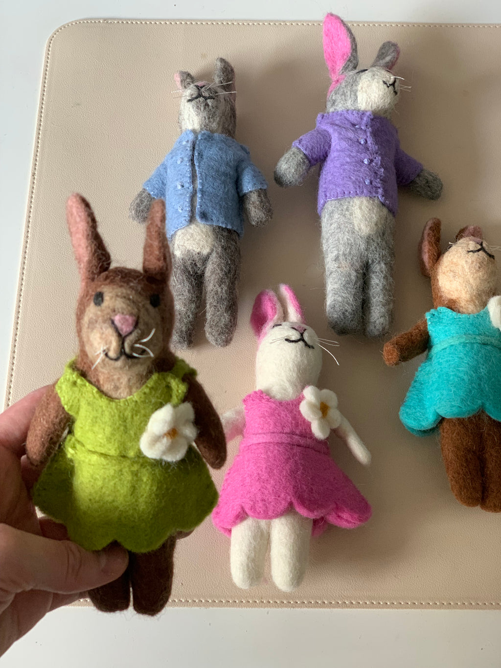 Five handmade felt animal dolls wearing colorful outfits, one held in hand on a light tan surface