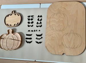 Wooden pumpkin-shaped trays and black eyebrows and mouths inserts arranged beside pumpkin outlines on wooden board on beige surface