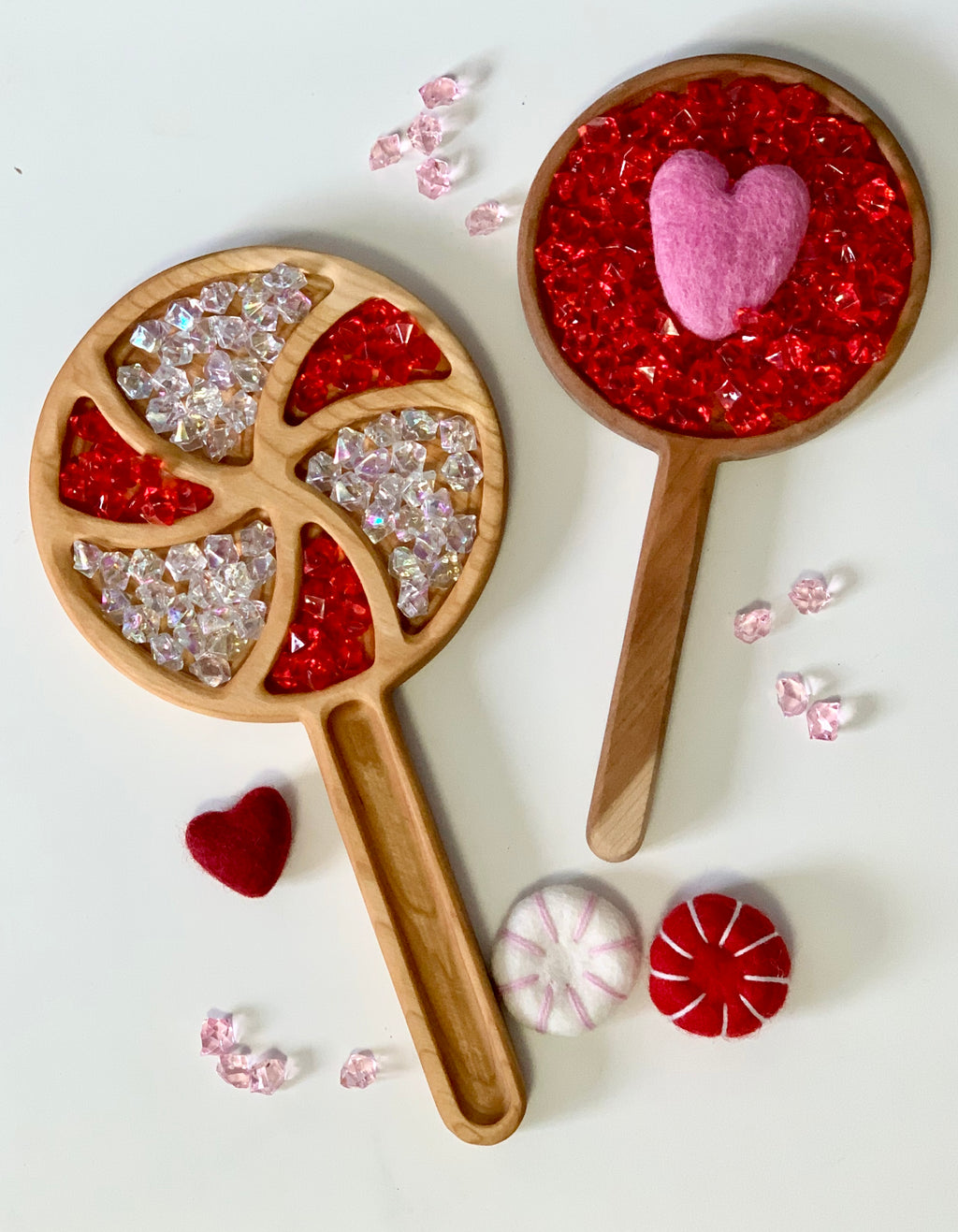 Wooden sensory trays shaped like lollipop and frying pan filled with red and clear gems and felt heart pieces on white surface