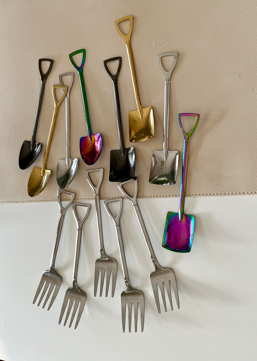 Collection of small metal shovels and forks in various metallic finishes, arranged on a light surface and fabric background