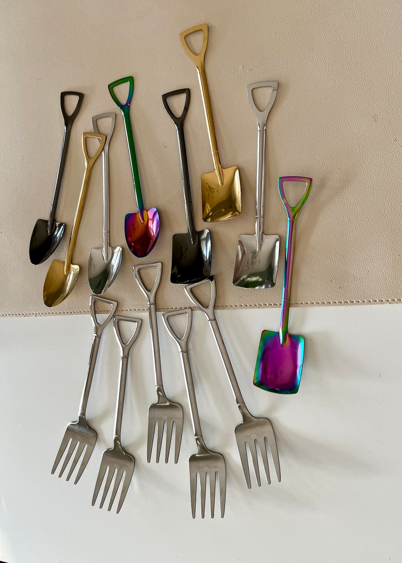 Collection of small metal shovels and forks in various metallic finishes, arranged on a light surface and fabric background