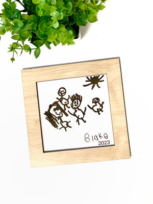 Child’s drawing traced in black ink on white paper, framed in natural wood square with green plant in background