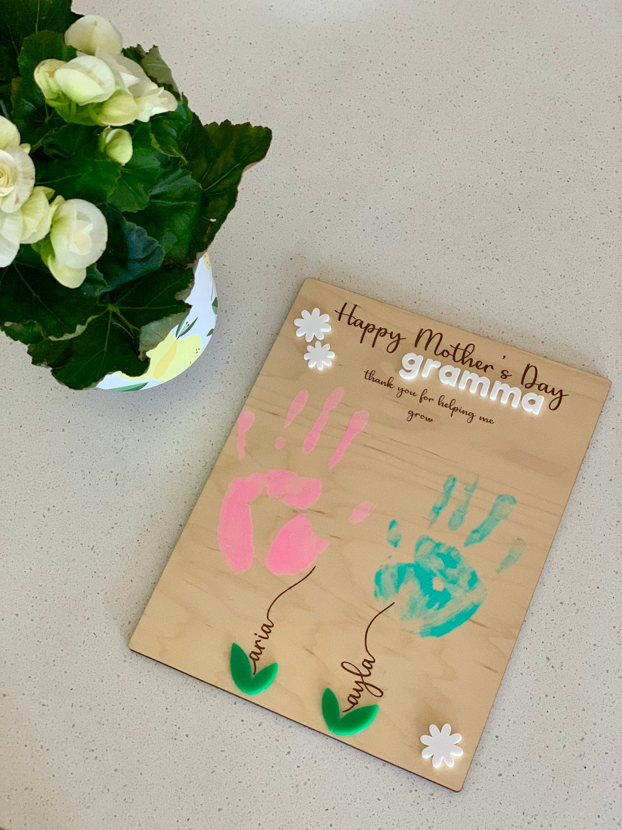 Wooden plaque with pink and blue handprints, green leaves, white flowers, and Mother's Day message for grandma on a beige surface