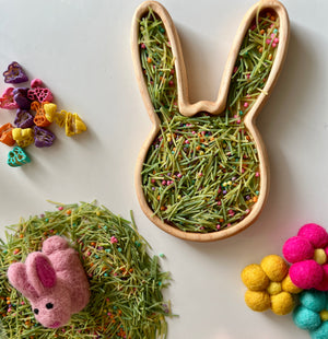 Bunny-shaped wooden tray filled with green pasta strands and colorful sprinkle pieces surrounded by felt flowers and shapes