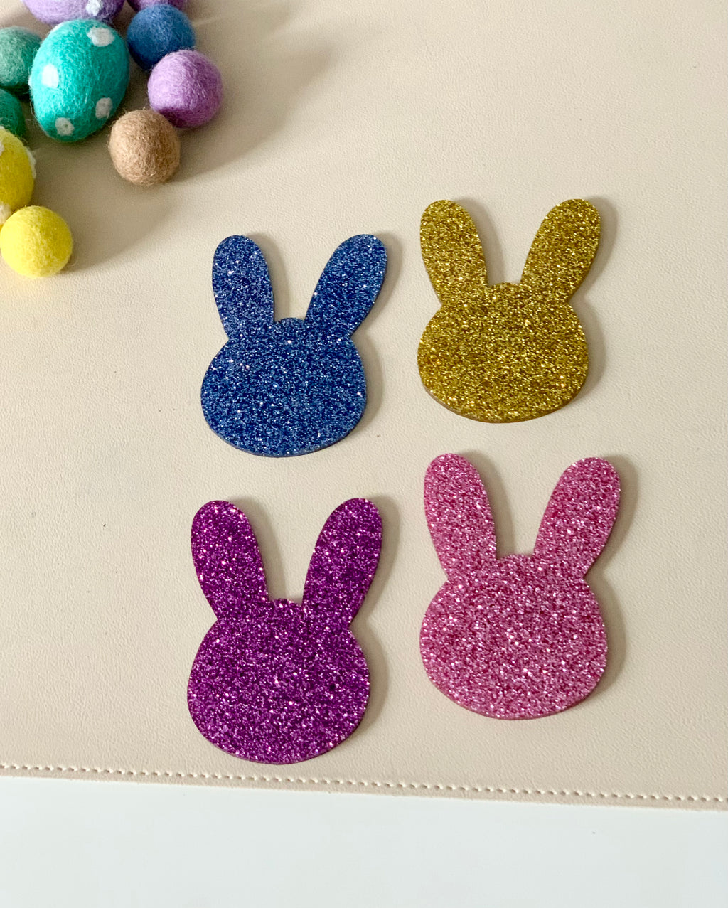 Four glittery bunny-shaped cutouts in blue, gold, purple, and pink on a light surface with felt balls nearby