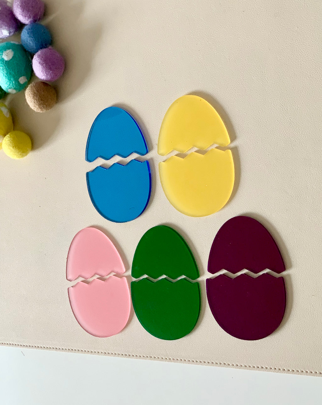 Five colorful cracked egg-shaped acrylic cutouts in blue, yellow, pink, green, and purple on a light surface with felt balls nearby