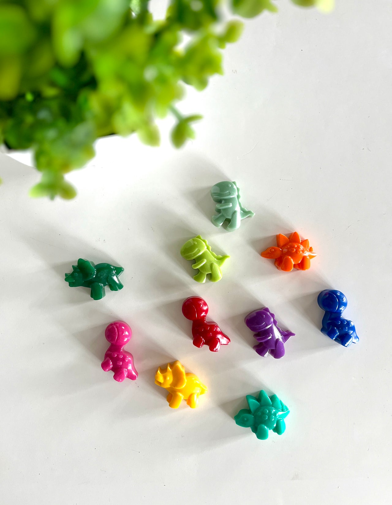 Ten small colorful dinosaur-shaped figures arranged on a white surface with a green plant blurred in the corner