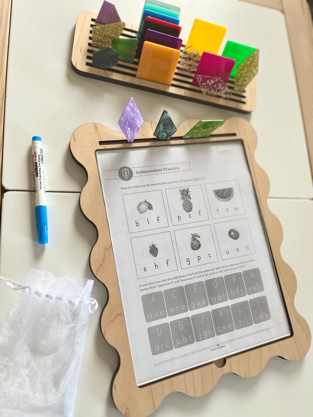 Wooden-framed activity board with scalloped edges holding a clear sheet over alphabet exercise and colorful translucent shape tiles above and dry erase marker nearby