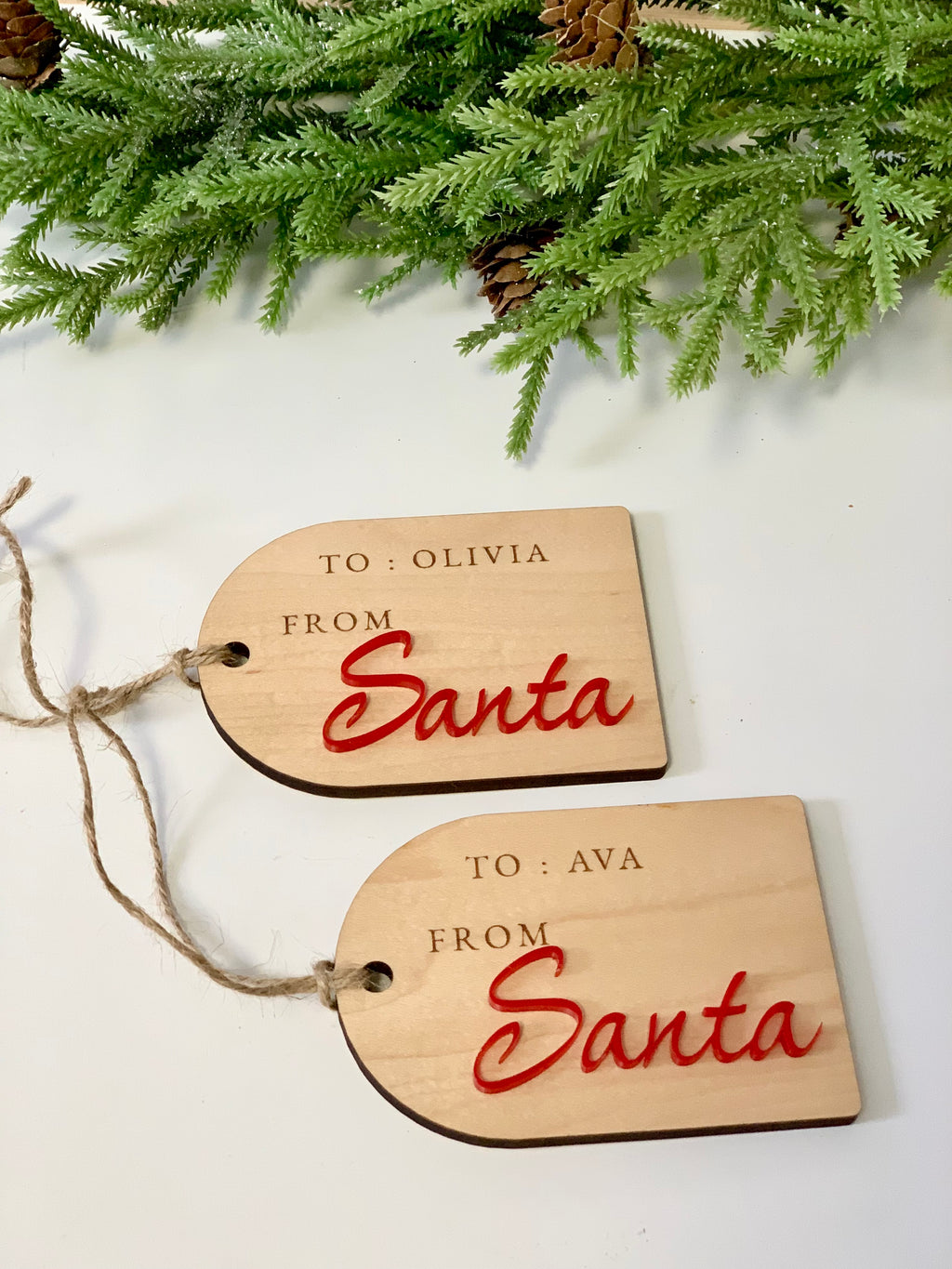 Two wooden gift tags with red "Santa" text and engraved recipient names on white surface with pine branches above