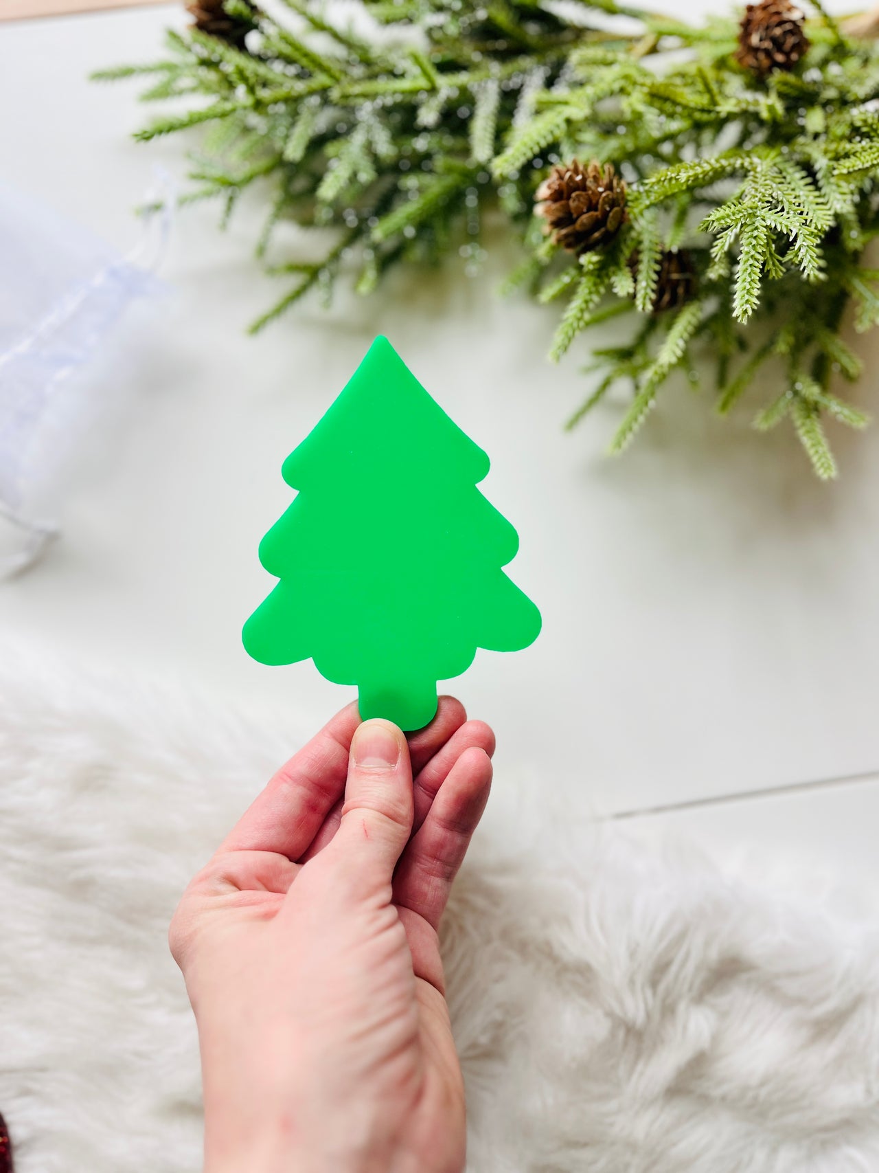 Hand holding small bright green Christmas tree-shaped cutout with pine branches and pinecones in background