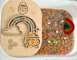 Wooden tray with rainbow and pot shapes filled with cereal rings beside a sensory bin of colorful rice and rainbow pieces