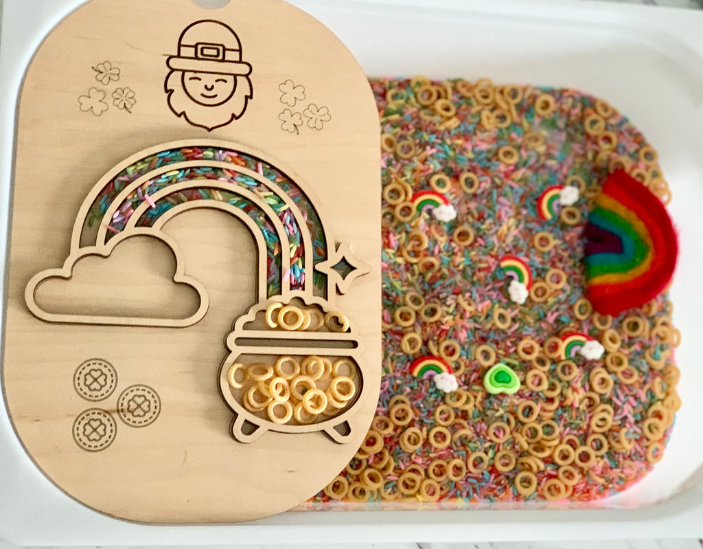 Wooden tray with rainbow and pot shapes filled with cereal rings beside a sensory bin of colorful rice and rainbow pieces