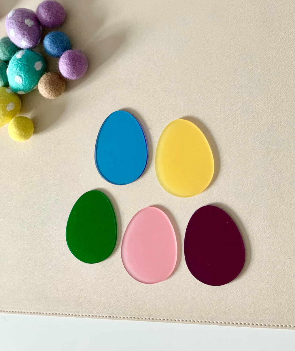 Five translucent egg-shaped pieces in blue, yellow, green, pink, and purple on a light surface next to felt balls
