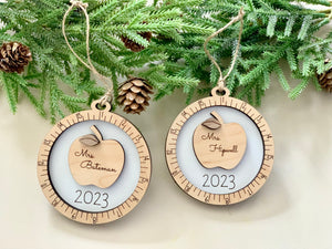 Pair of personalized wooden teacher Christmas ornaments with apple design and 2023 inscription hanging from twine on greenery background
