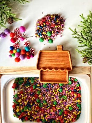 Wooden house-shaped dish with assorted colorful beads, felt balls, peppermint candies, and greenery with pine cones on white surface.