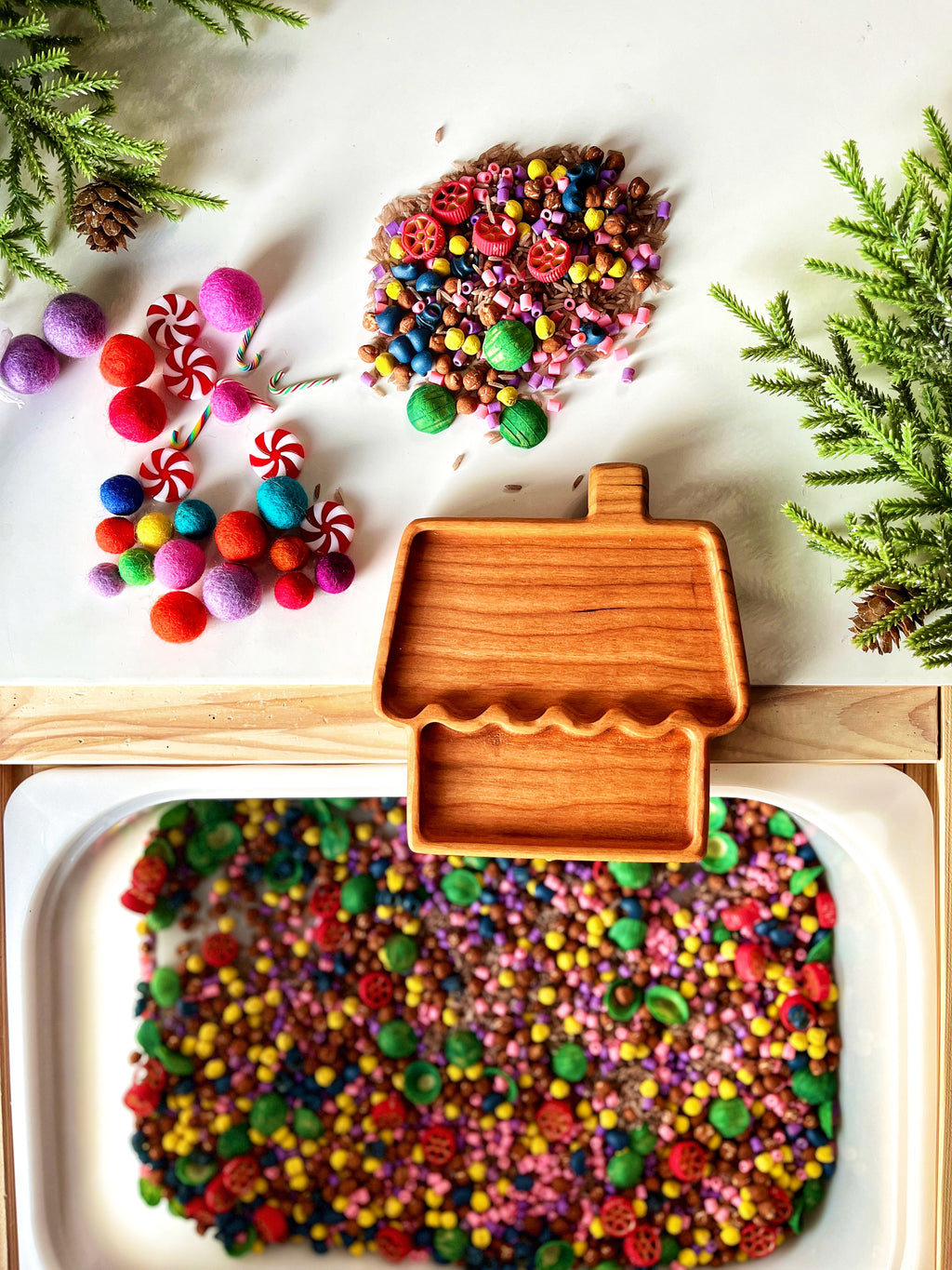 Wooden house-shaped dish with assorted colorful beads, felt balls, peppermint candies, and greenery with pine cones on white surface.