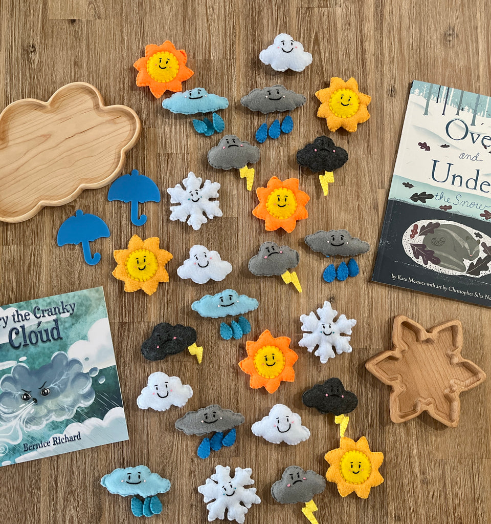 Colorful felt weather shapes including suns, clouds, rain, snowflakes, umbrellas, and lightning bolts arranged on a wooden surface with two wooden trays and children's books, featuring Felt Weather Shapes, Made in Canada, handcrafted, homeschool, education resources.