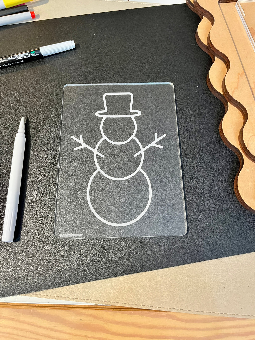 Clear plastic board with an outlined snowman wearing a top hat and stick arms on a black surface next to markers