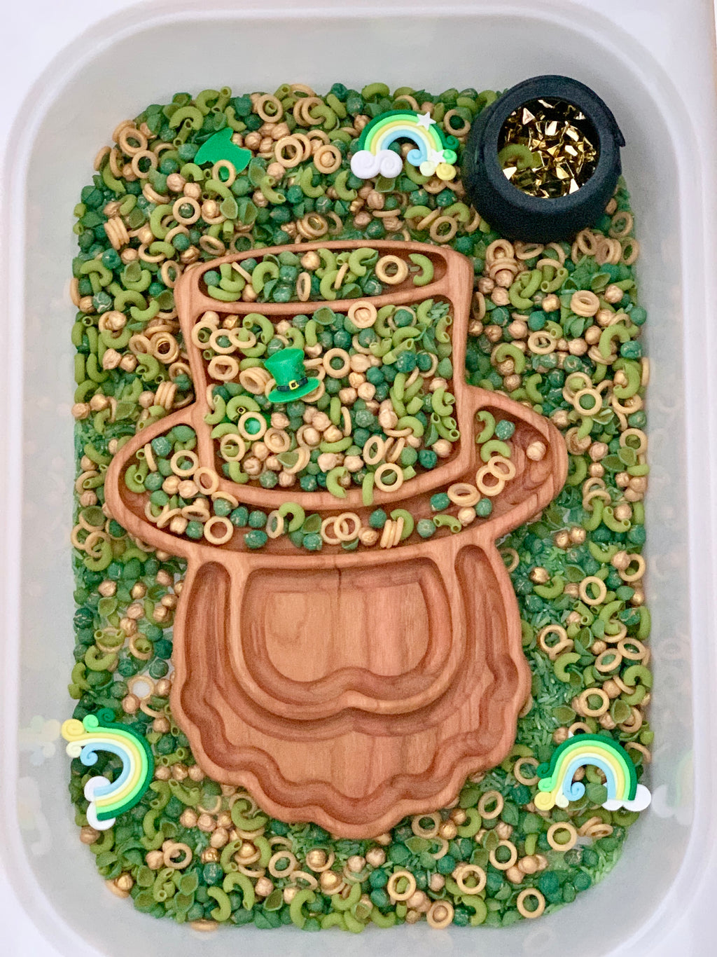 Plastic bin filled with green and beige pasta shapes, wooden leprechaun tray, rainbows, and a small black pot with gold stars