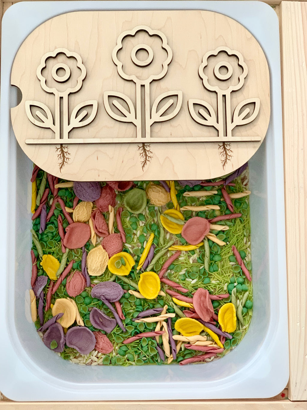 White rectangular sensory bin filled with colorful dried pasta and flower-themed wooden puzzle lid on top