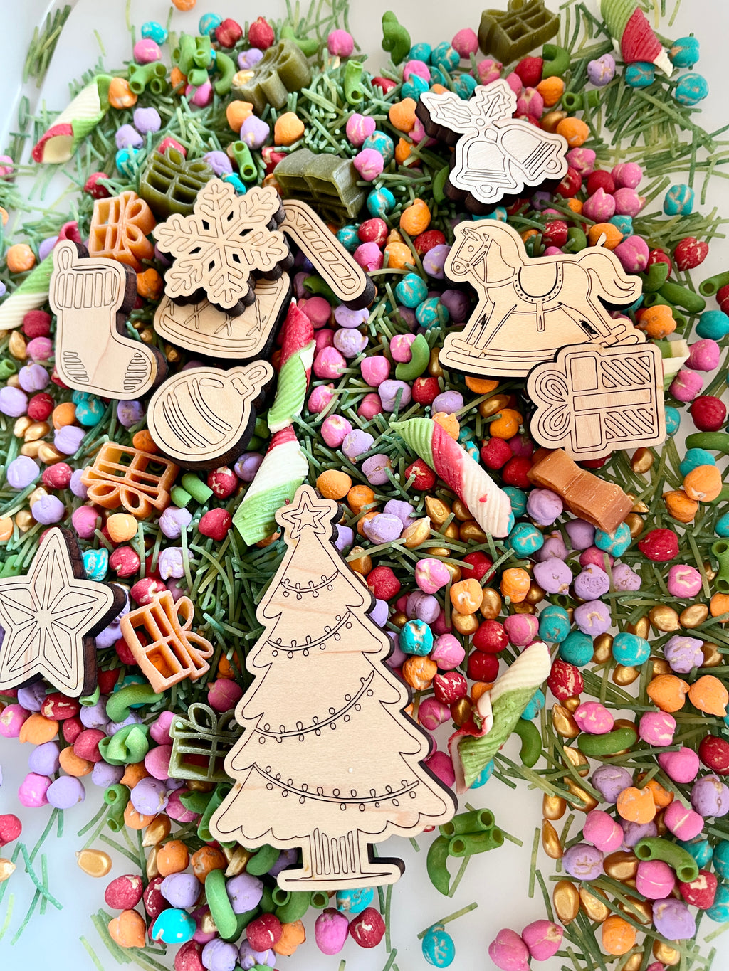 Assortment of colorful small beads, green dried botanicals, striped pasta, and wooden Christmas-themed cutouts on white surface