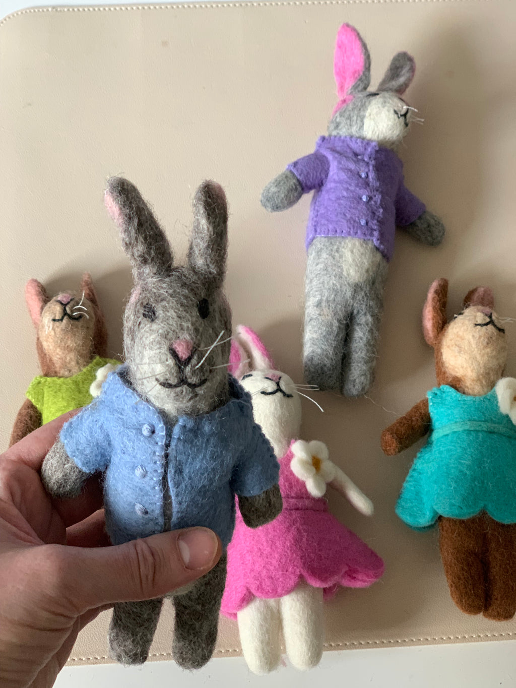 Set of five needle-felted bunnies dressed in colorful handmade felt clothes on a beige surface with one held in hand