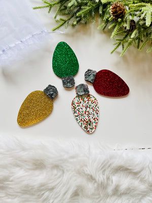 Four glittery acrylic Christmas light bulb shapes in red, green, gold, and confetti pattern on white surface with greenery nearby
