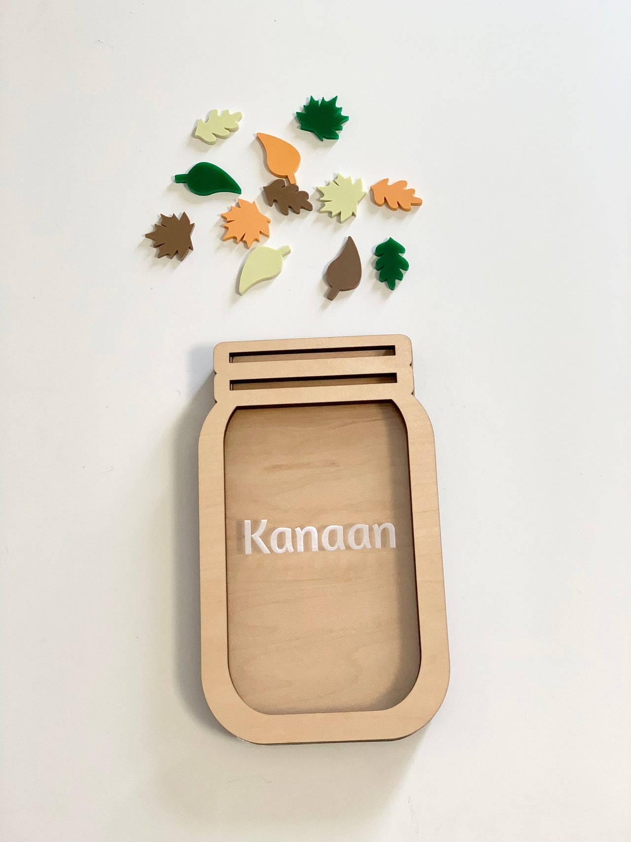 Light wooden jar-shaped frame with the name Kanaan and multicolored wooden leaf tokens scattered above on a white background
