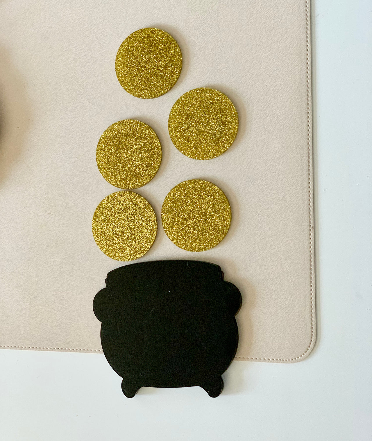 Black pot-shaped cutout with five gold glittery circular pieces arranged above on beige surface