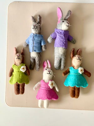 Five handmade felt bunny dolls dressed in colorful clothes arranged on a light beige surface