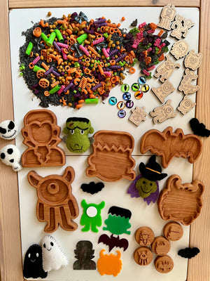 Halloween-themed wooden trays, colorful pasta shapes, felt ghosts, monsters, and decorative craft stamps on wooden table.