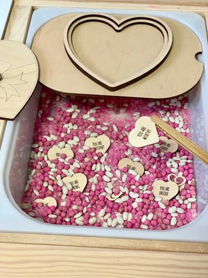 Sensory bin with pink and white beans, small wooden hearts engraved with positive messages, and a heart-shaped wooden lid.