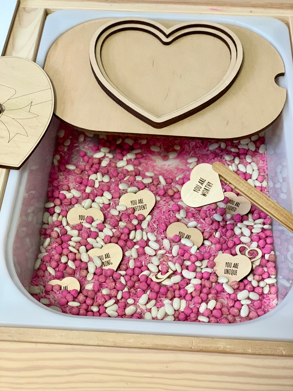 Sensory bin with pink and white beans, small wooden hearts engraved with positive messages, and a heart-shaped wooden lid.