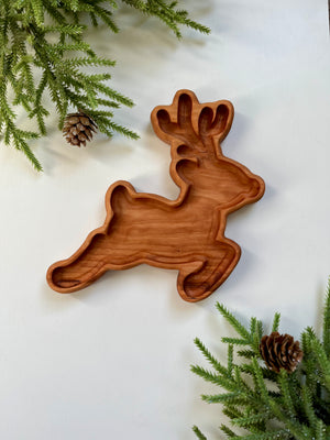 Wooden tray carved in the shape of a reindeer surrounded by green pine branches and pine cones on white surface