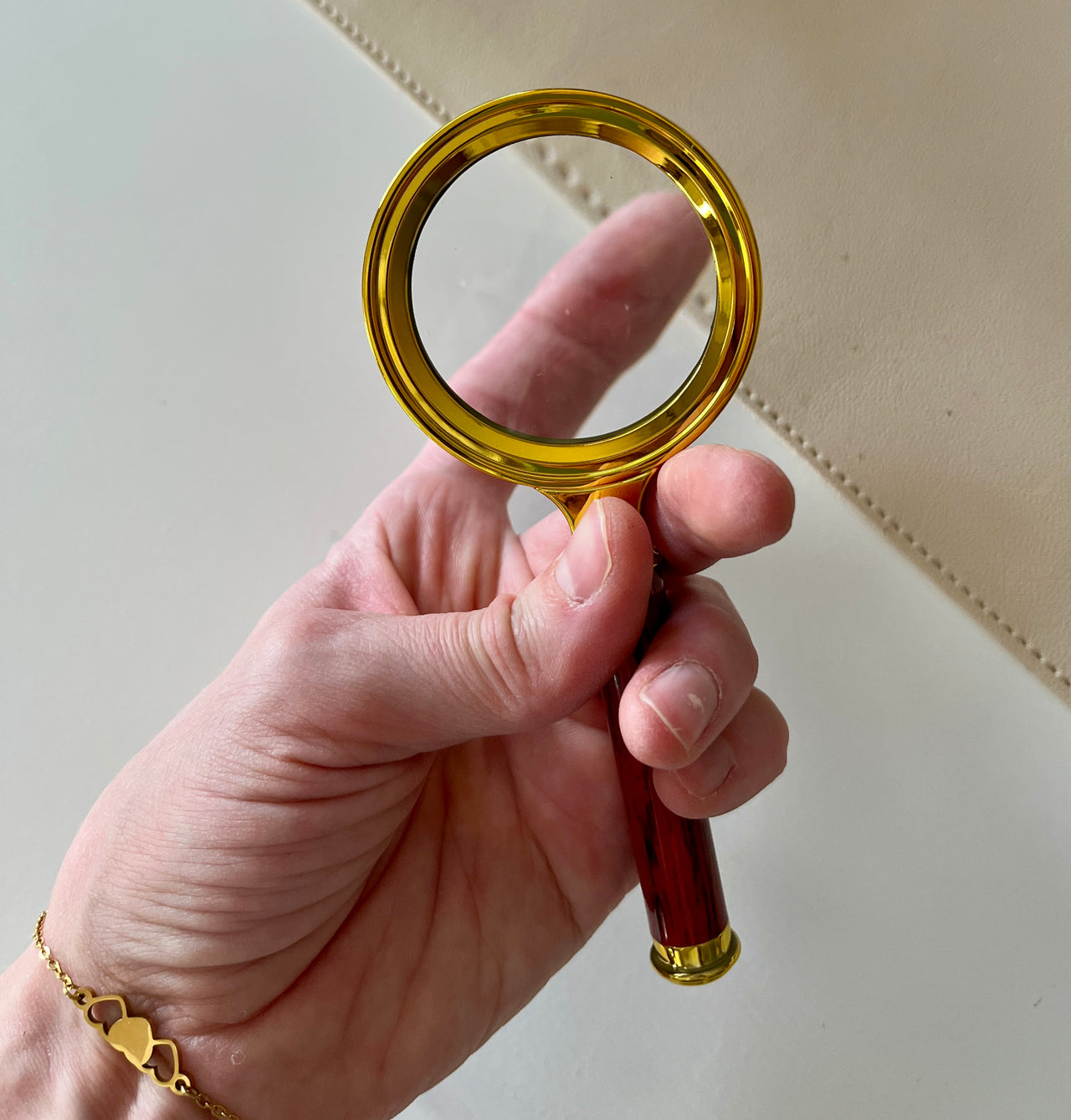 Hand holding a magnifying glass with a gold rim and dark wooden handle over a light neutral background