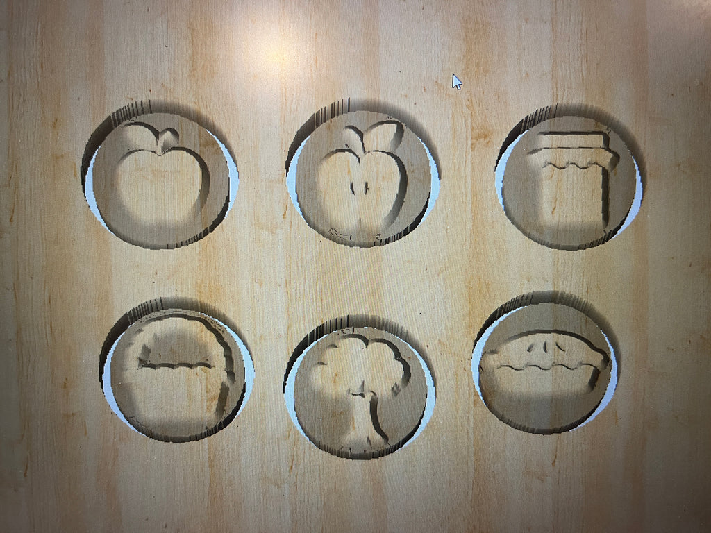 Wooden board with six carved shapes including apple, peach, jam jar, pie slice, tree, and pie on light wood surface