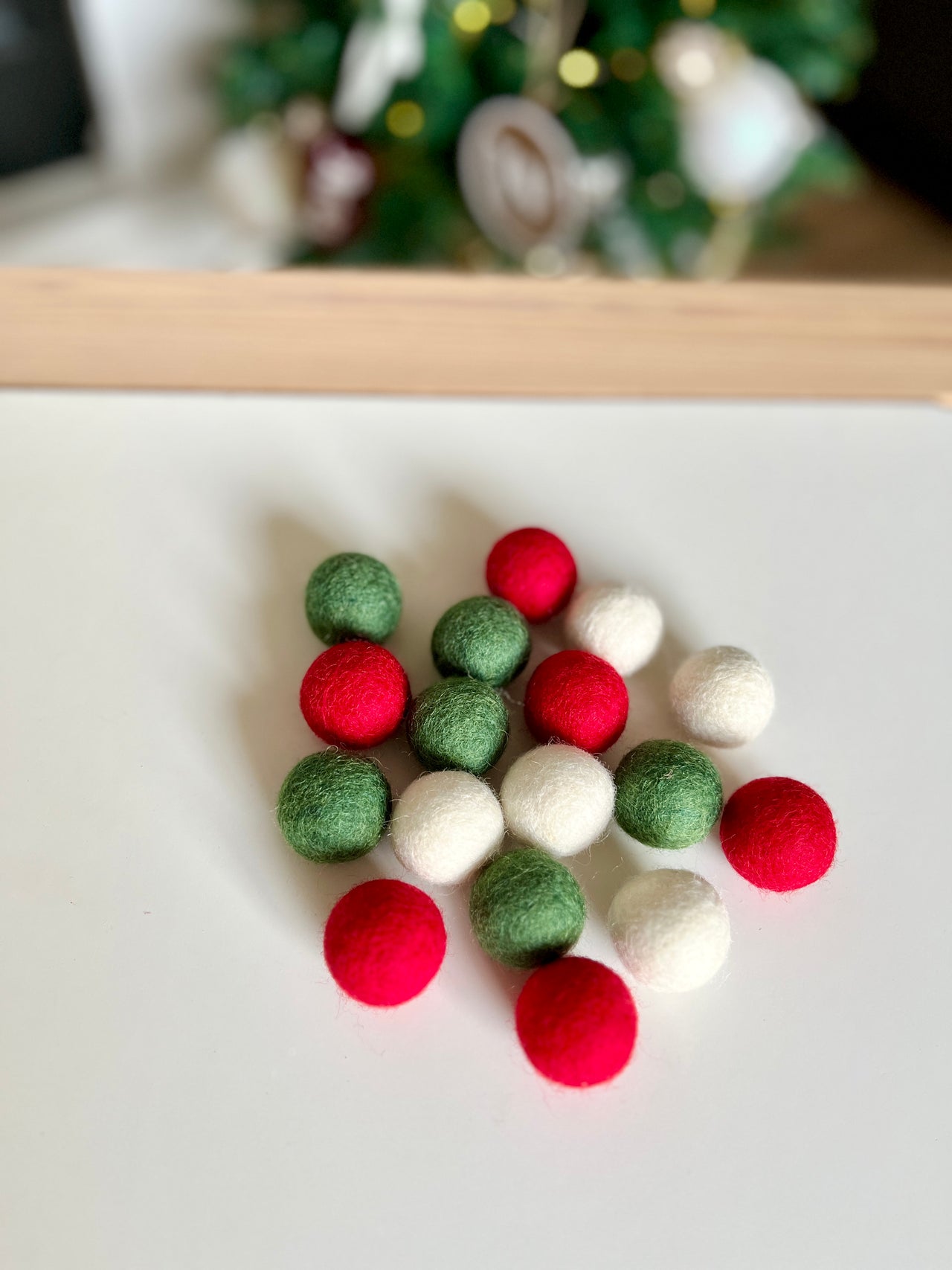 Small red, green, and white felt balls arranged on a white surface with blurred holiday decorations in the background