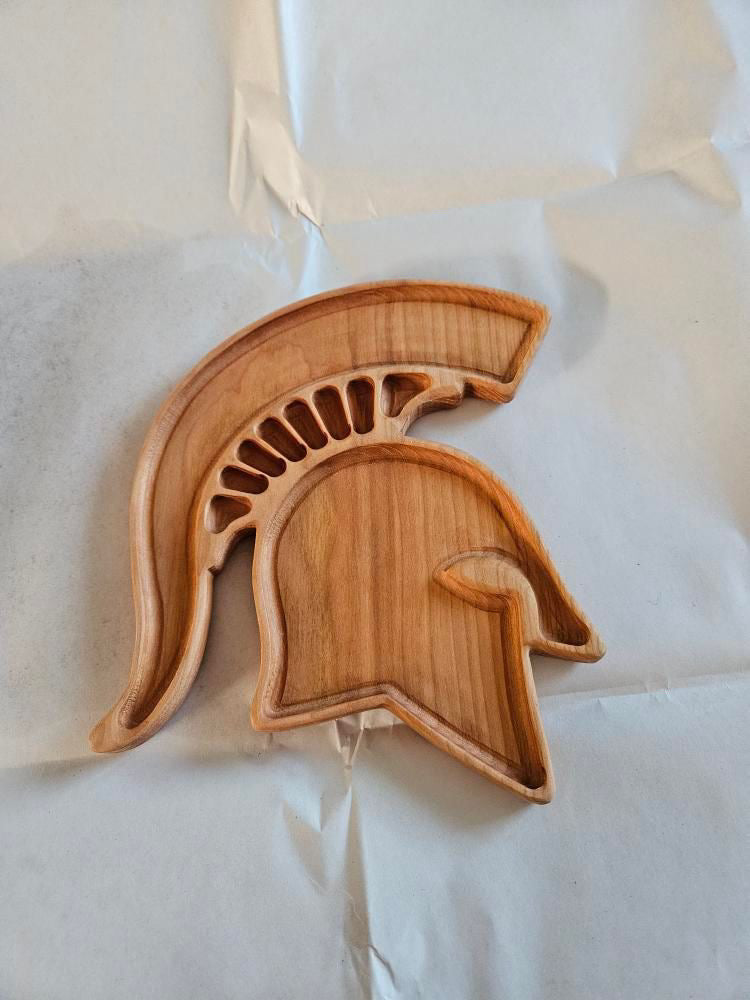 Wooden tray carved in the shape of a Spartan helmet with detailed cutouts on a light fabric background