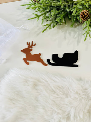 Brown reindeer and black sleigh acrylic cutouts placed on a white surface with greenery and pinecone nearby
